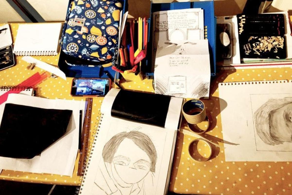 Photograph of art materials on a table and an open sketchbook with drawing of a portrait
